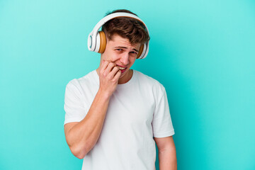 Young caucasian man listening to music with headphones isolated on blue background biting...