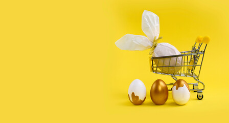 Mock up Easter composition with shopping trolley,  golden painted and bunny wrapped eggs on yellow background. Minimalist concept for holiday or sale. Banner. Copyspace.