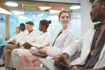 Multi-ethnic group of young people wearing lab coats and smiling while sitting in row and during...