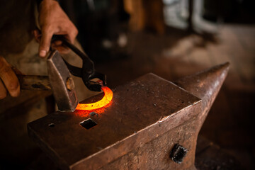 The blacksmith forging the molten metal on anvil in smithy. Blacksmith at the workshop. Working metal with hammer and tools in forge - obrazy, fototapety, plakaty