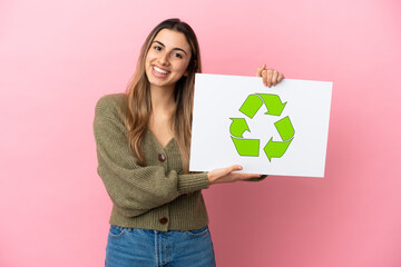 Young caucasian woman isolated on pink background holding a placard with recycle icon with happy expression