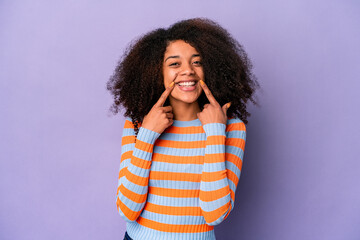 Young african american curly woman isolated on purple background smiles, pointing fingers at mouth.