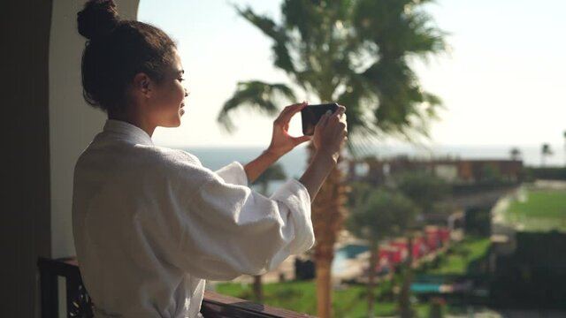 Young woman in bathrobe taking pictures of beautiful tropical landscape and posting it on social network then enjoying the view, standing on hotel balcony. Vacation, technology concept