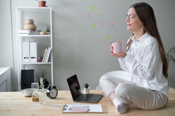 Young smiling business woman sits in lotus position desktop in front of laptop and holds mug in her hands.