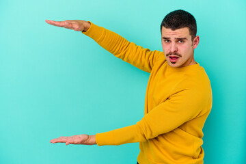 Young caucasian man isolated on blue background shocked and amazed holding a copy space between hands.