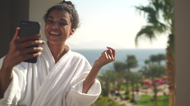 Excited young woman in bathrobe communicating with her friends and colleagues via video chat app, telling about her tropical vacation, standing on hotel balcony. Resort, technology concept