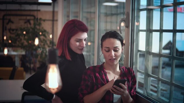 Two woman discuss new smartphone app design standing next to coworking office window