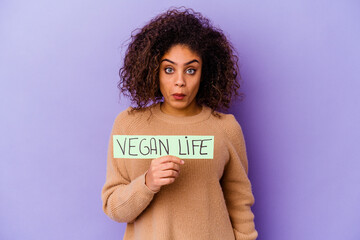 Young African American woman holding a Vegan life placard isolated shrugs shoulders and open eyes confused.