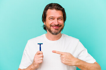 Middle age dutch man holding a razor blade isolated on blue background smiling and pointing aside, showing something at blank space.