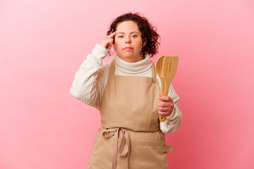 Woman with Down syndrome cooking at home isolated on pink background pointing temple with finger, thinking, focused on a task.