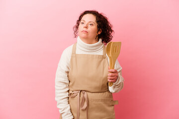 Woman with Down syndrome cooking at home isolated on pink background confused, feels doubtful and unsure.