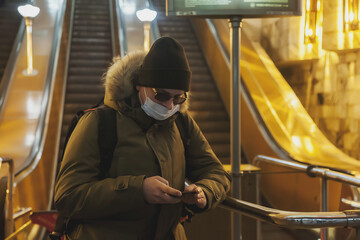 Young man in wear casual winter clothes with glasses and medical mask at metro escalator. Attractive teenager with phone in big city subway, looking at mobile phone and emotions of an unexpected email