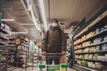 Young man in wear casual winter clothes with glasses and medical mask с grocery cart in hypermarket. Attractive teenager with backpack in city shop, looking at camera and emotions of an unexpected