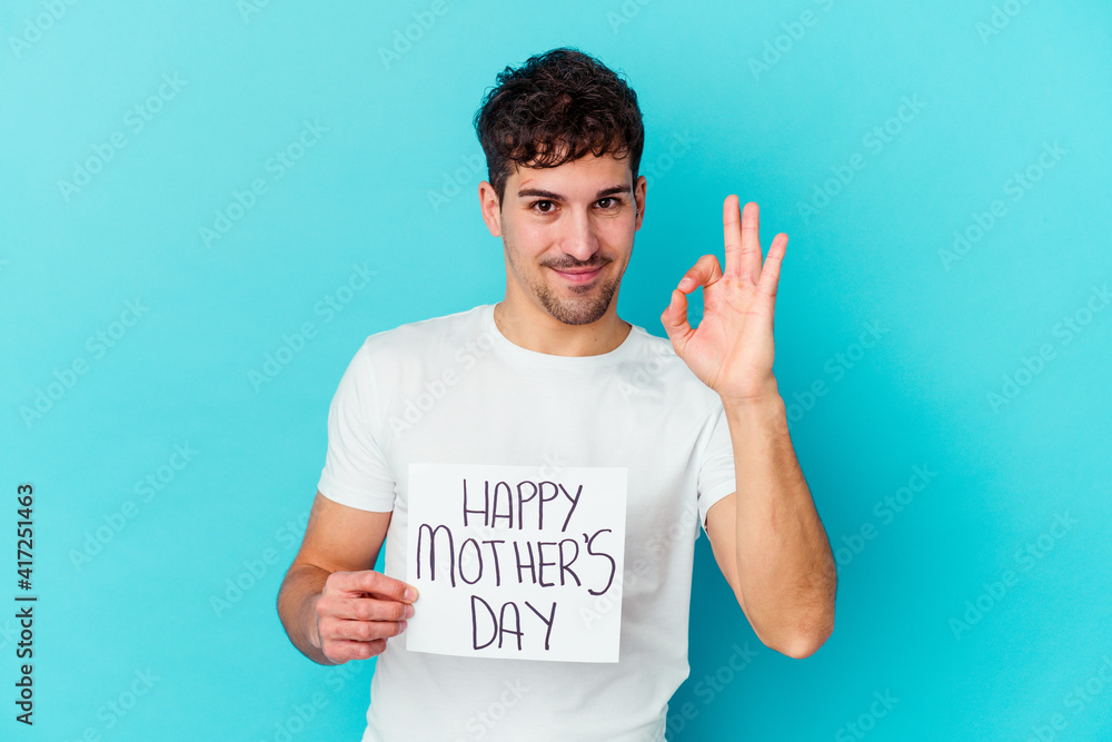 Wall mural Young caucasian man holding a happy mothers day placard isolated cheerful and confident showing ok gesture.