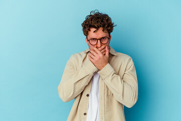 Young caucasian man wearing eyeglasses isolated on blue background covering mouth with hands looking worried.