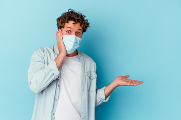 Young caucasian man wearing an antiviral mask isolated on blue background holds copy space on a palm, keep hand over cheek. Amazed and delighted.