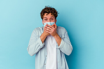 Young caucasian man wearing an antiviral mask isolated on blue background shocked covering mouth with hands.