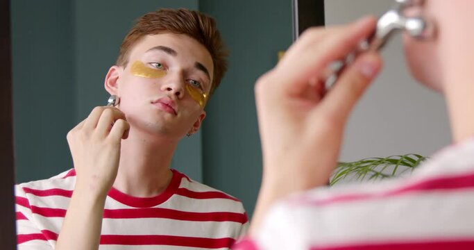 Young man makes face massage with a massager roller in front of a mirror at home. Facial skin care lifting and beauty rituals concept.