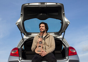 Smiled young man with ukulele in his hands sitting in the trunk. Young man playing ukulele