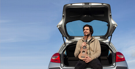Smiled young man with ukulele in his hands sitting in the trunk. Young man playing ukulele. Copy space