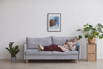 Wide angle view at modern mixed-race woman using smartphone while lying on sofa in minimal home interior, copy space