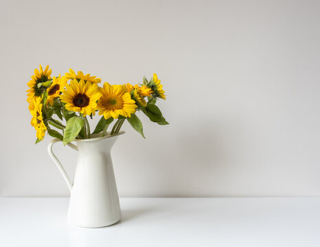 Close up of sunflowers in white jug on shelf against neutral wall background (selective focus)