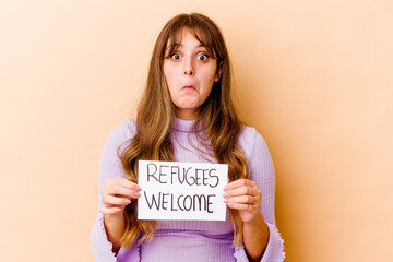 Young caucasian woman holding a Refugees welcome placard isolated shrugs shoulders and open eyes...