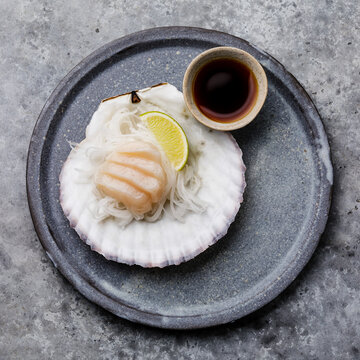 Live scallop sashimi on shell with daikon, lime and soy sauce on gray background