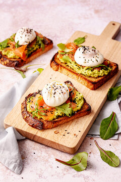 3 smoked salmon toasts with avocado cream, poached eggs and sesam seeds on top, served on a cutting board.