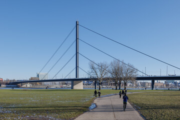 Outdoor sunny view of people walk on street along natural riverside of Rhine River, and background cityscape and suspension bridge in winter season in Düsseldorf, Germany.