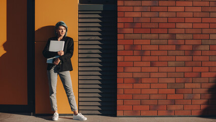 Charming lady with blue hair posing on a brick wall in a sunny day embracing a computer