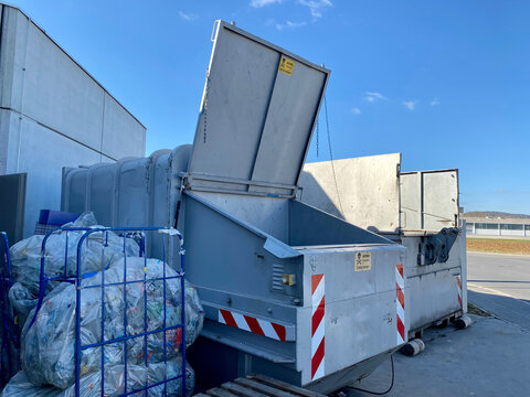 Grauer Container, ein Fl&uuml;gel weit ge&ouml;ffnet f&uuml;r plalttgepresste Pfandplastikflaschen in Plastikbeutel verpackt. Betonwand und strahlend blauer Himmel drum herum.