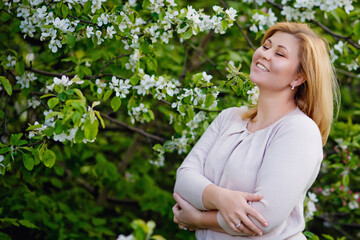 Portrait of a fat plump young blonde woman blooming with an apple tree on a white floral background in a park on a spring day. Happy plus-size girl with her eyes closed enjoying spring.