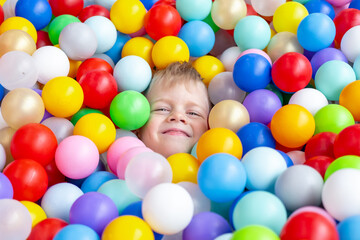 Fototapeta na wymiar Blonde little boy lying on multi coloured plastic balls in big dry paddling pool in playing centre. Smiling at camera. Portrait close up. Having fun in playroom. Leisure Activity.