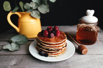 Juicy pancakes with berries and honey on a white plate, spoon, jar, wooden table