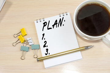 The word plan is written on a notepad that sits on a wooden table next to a white coffee cup and stationery.