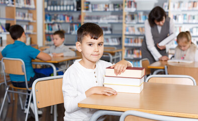 Fototapeta premium Portrait of tired preteen boy sitting at desk with pile of books in classroom..