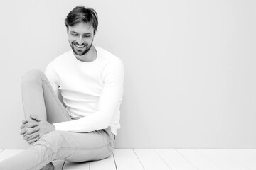 Portrait of handsome smiling hipster lumbersexual businessman model wearing casual white sweater and trousers. Fashion stylish man sitting against light blue wall in studio