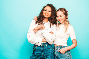 Two young beautiful smiling international hipster female in trendy summer clothes. Sexy carefree women posing near blue wall in studio. Positive models having fun. Concept of friendship