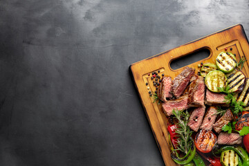 Sliced fried medium steak with grilled vegetables on wooden cutting board on dark background with copy space top view