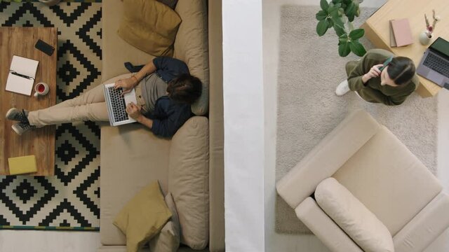 Top view shot of young man sitting on couch and working on laptop in his apartment