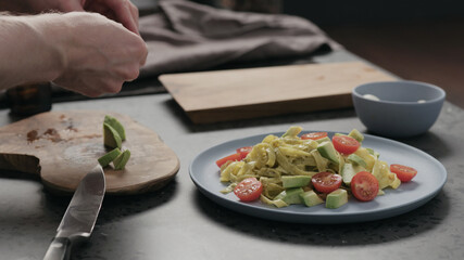 man add cherry tomatoes to pesto fettuccine on blue plate