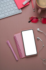 Above view of female workspace with smart phone, earphone, notebook and keyboard on pastel pink background.