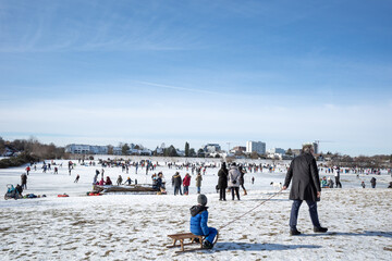 Outdoor sunny view, crowd of people enjoy outdoor activities, playing pond hockey, sledding ice skating on the snow on riverside of Rhine River in winter season in Düsseldorf, Germany.