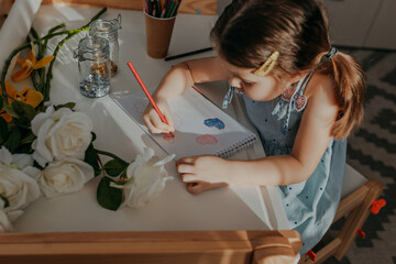 Fototapeta na wymiar Little girl drawing hearts on white paper. Happy mother's day card with girl sitting near white table. Kid makes homework with pencils. Girl with flowers.