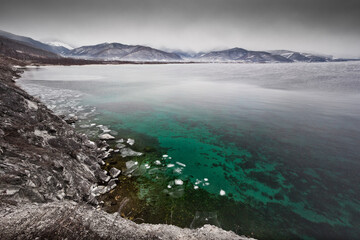 Icy coast, green clear water and mountains in the distance, Lake Baikal in late autumn