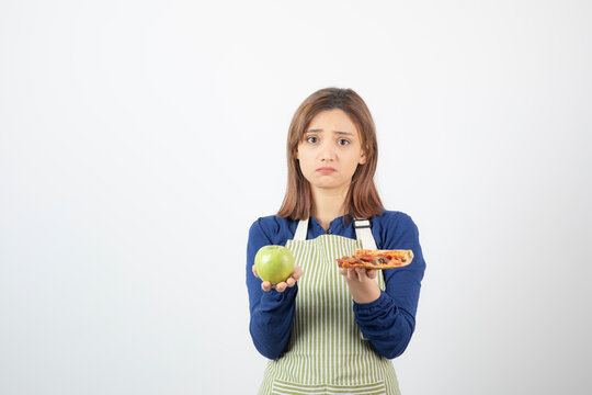 Image of woman in apron trying to choose what to eat apple or pizza