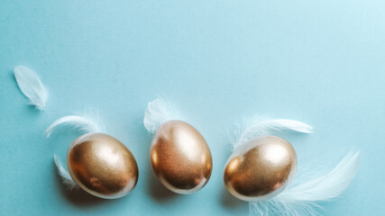Golden easter colour eggs with white feathers on pastel blue background in Happy Easter decoration. Spring holiday top view concept.