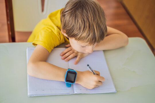Little boy sitting at the table and looking smart watch. Smart watch for baby safety. The child makes school lessons, listening to music, calling friends