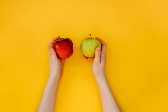 Female hands holding two apples, red and green, on yellow background.